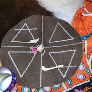 May include: A brown circular felt mat with four white triangles drawn on it. The triangles are divided by a white cross. There are small white bones and pink and white figurines on the mat.