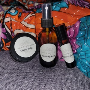 May include: Three small bottles with labels that read "Clarity Balm", "Cleansing Spray", and "Agua de Flor". The bottles are on a patterned fabric background.