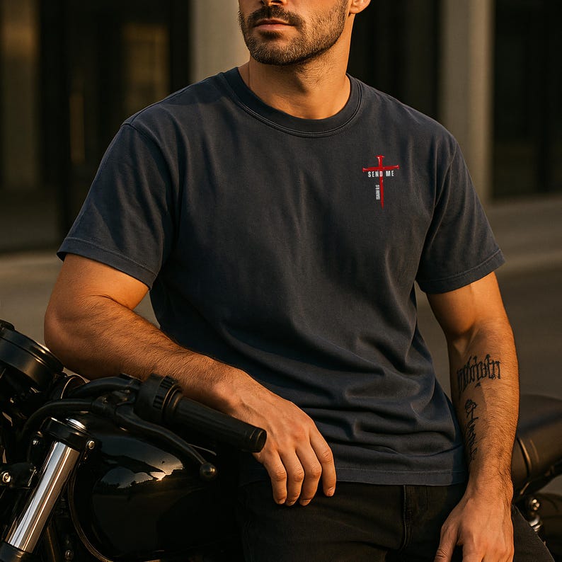 May include: A dark charcoal gray short-sleeve t-shirt featuring a small graphic on the upper left chest. The graphic displays a red cross with the text "SEND ME" in white capital letters positioned horizontally across the center of the cross.
