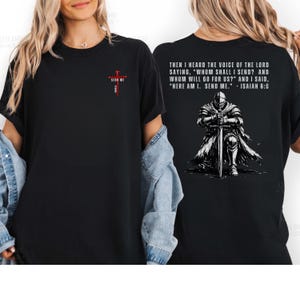 May include: A black t-shirt featuring a small red cross graphic on the front chest with the text SEND ME. The back displays a graphic of a knight kneeling with a sword and the text: THEN I HEARD THE VOICE OF THE LORD SAYING, WHOM SHALL I SEND?