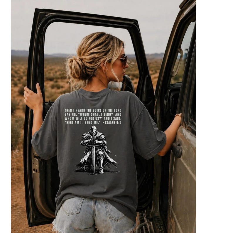 May include: A charcoal gray short-sleeve t-shirt featuring a graphic of a knight kneeling with a sword. The text reads: THEN I HEARD THE VOICE OF THE LORD SAYING, "WHOM SHALL I SEND? AND WHOM WILL GO FOR US?" AND I SAID, "HERE AM I. SEND ME." - ISAIAH 6:8