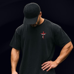 May include: A black baseball cap and a black short-sleeve t-shirt. The shirt features a small graphic on the upper left chest consisting of a red cross made of nails with the text "SEND ME" in white letters above the vertical bar of the cross.