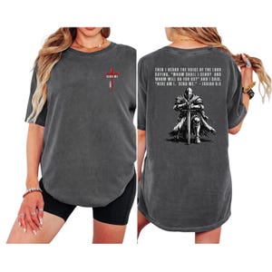 May include: A charcoal gray short-sleeve t-shirt featuring a small red cross graphic on the front chest with the text "SEND ME". The back displays a graphic of a knight kneeling with a sword and the text "THEN I HEARD THE VOICE OF THE LORD..."