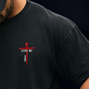 May include: A black t-shirt featuring a graphic on the chest. The design consists of a red cross made of nails with the white text SEND ME across the horizontal bar and the white text ISAIAH 6:8 printed vertically along the bottom of the cross.