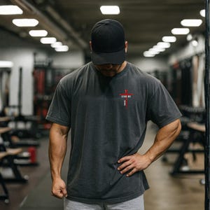 May include: A charcoal gray short-sleeve t-shirt featuring a small red cross graphic on the left chest with the text "SEND ME" printed in white letters vertically along the cross. The shirt has a crew neckline and a relaxed, boxy fit.