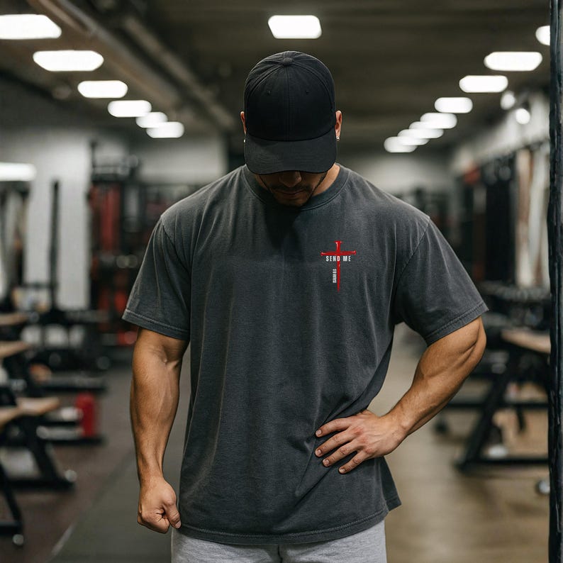 May include: A charcoal gray short-sleeve t-shirt featuring a small red cross graphic on the left chest with the text "SEND ME" printed in white letters vertically along the cross. The shirt has a crew neckline and a relaxed, boxy fit.