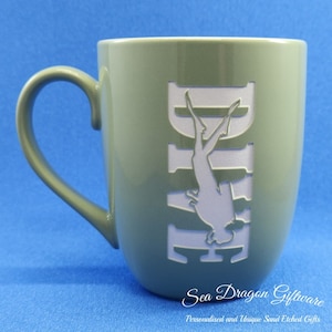 May include: A green ceramic mug with a white silhouette of a scuba diver etched into the side. The mug has a rounded shape and a handle. The text "Sea Dragon Giftware" and "Personalised and Unique Sand Etched Gifts" is printed on the bottom of the mug.