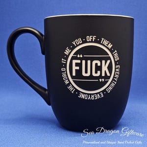 May include: Black ceramic mug with a white interior and a large handle. The mug features the word "FUCK" in a white circle, surrounded by the phrases "ME, YOU, OFF, THEM, THIS, EVERYTHING, EVERYONE, THE WORLD, IT".