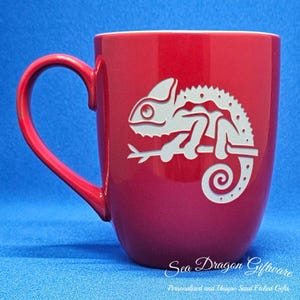 May include: A red ceramic mug featuring a white chameleon design. The mug has a rounded base and a handle. The text "Sea Dragon Giftware" is printed on the bottom of the mug.
