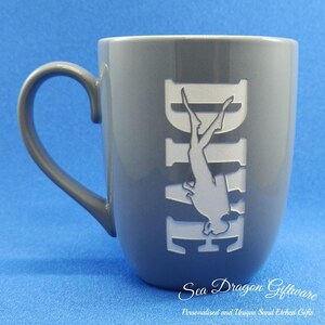 May include: Grey ceramic mug with a white silhouette of a scuba diver etched into the side. The mug reads "Dive" in white lettering. The mug is on a blue surface.