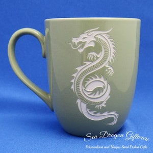 May include: A green ceramic mug with a white sand-etched dragon design. The mug has a rounded body and a handle. The text "Sea Dragon Giftware" and "Personalised and Unique Sand Etched Gifts" is printed on the bottom of the mug.