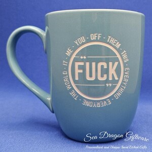 May include: A blue ceramic mug with a curved handle. The mug features the word "FUCK" in a circular design, surrounded by the phrases "ME - YOU - OFF - THEM - THIS - EVERYTHING - EVERYONE - THE WORLD - IT". The mug has a white interior.
