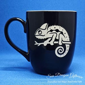 May include: Black ceramic mug featuring a white chameleon design. The mug has a rounded shape and a handle. The text "Sea Dragon Giftware" is printed on the bottom of the mug.