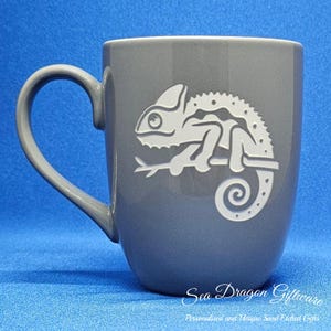 May include: Grey ceramic mug featuring a white chameleon design. The mug has a rounded shape and a handle. The chameleon is facing right and has a curled tail. The text "Sea Dragon Giftware" is printed on the bottom of the mug.