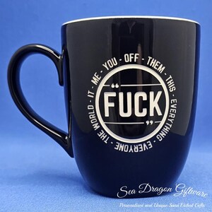May include: Black ceramic mug with a white interior and handle. The mug features the word "FUCK" in a white circle, surrounded by text. The mug also has the text "Sea Dragon Giftware" at the bottom.
