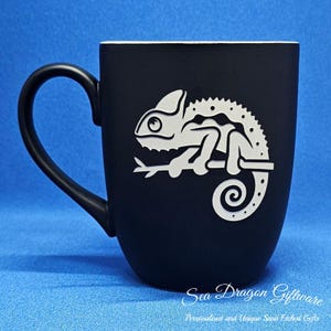 May include: Black ceramic mug with a white silhouette of a chameleon on a branch. The mug has a white rim and handle. The text "Sea Dragon Giftware" is printed on the bottom of the mug.