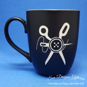 May include: Black ceramic mug featuring a white design of a button, needle, and scissors. The text "Sea Dragon Giftware" and "Personalized and Unique Sand Etched Gifts" is at the bottom of the mug.