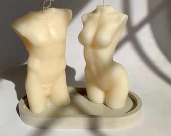 May include: Two white, sculpted candles in the shape of a male and female torso, sitting on a light gray oval tray.