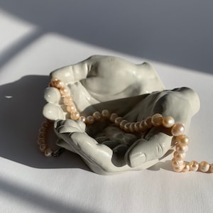 May include: A white ceramic sculpture of two hands cupped together, holding a strand of pink pearls.