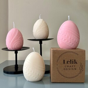 May include: Four egg-shaped candles in pink and cream, with detailed embossed patterns. Two candles are on black metal stands, one on a cardboard box with the text "Lelik CRAFT DESIGN".