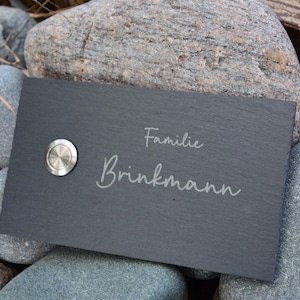Op de afbeelding: Een rechthoekig, donkergrijs leistenen naambord met de woorden "Familie Brinkmann" in een cursief lettertype gegraveerd. Een zilveren deurbelknop bevindt zich aan de linkerkant. Het bord rust op grijze rotsen.