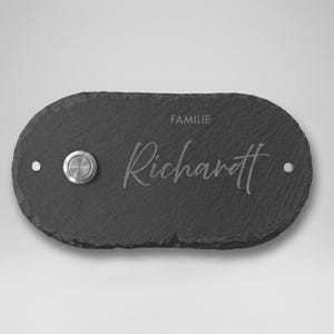 Könnte beinhalten: Ein dunkelgraues, ovales Klingelschild aus Schiefer mit dem Wort "FAMILIE" über dem Namen "Richardt" in einer Schreibschrift. Auf der linken Seite befindet sich ein silberner Klingelknopf, mit einem kleinen silbernen Punkt auf jeder Seite.