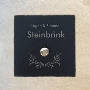 Könnte beinhalten: Schwarzes Schiefer-Türschild mit dem Text "Jürgen & Simone Steinbrink" und einem silbernen Türklingelknopf mit einem dekorativen Kranz-Design.