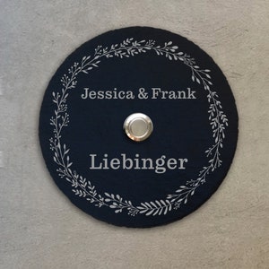 Könnte beinhalten: Schwarzes Schiefer-Türschild mit einem silbernen Türklingelknopf. Das Schild hat ein weißes Blumenkranz-Design und den Text "Jessica & Frank Liebinger" in der Mitte eingraviert.