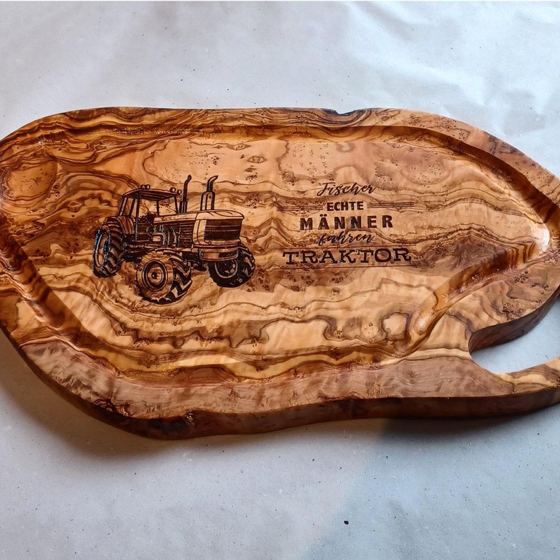 Olivenholzbrett mit Grifflasche mit natürlicher Holzmaserung. Das Brett hat ein geschnitztes Design eines Traktors und den Text "Fischer Echte Männer Fahren Traktor".