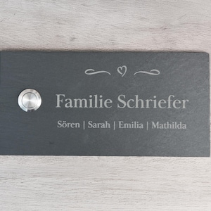 Peut inclure: Une plaque de porte rectangulaire en ardoise gris foncé avec un bouton de sonnette argenté à gauche. La plaque porte le nom de famille "Familie Schriefer" et les noms "Sören | Sarah | Emilia | Mathilda" en gris clair.