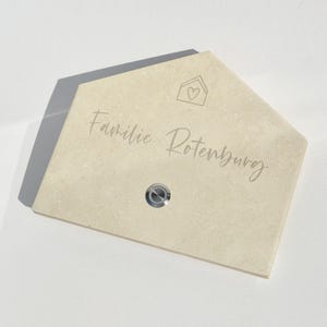 May include: A beige, house-shaped stone plaque with the engraved text "Familie Rotenburg" and a small house icon with a heart. A silver doorbell button is set into the stone.