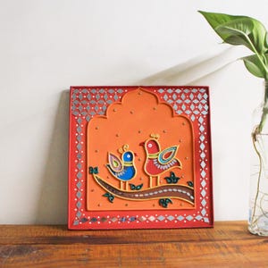 Puede incluir: Una obra de arte cuadrada y enmarcada con dos pájaros estilizados posados en una rama. Los pájaros son rojos y azules con diseños decorativos. El marco es rojo con detalles de espejo, sobre un fondo naranja.