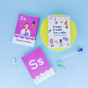 May include: Two pink spiral bound flip books with the letter "S" and the words "Space Scientist" on the cover. The book on the left has an illustration of a female space scientist. The book on the right has the words "Dream, Create, Innovate. A to Z of STEM careers flip book".