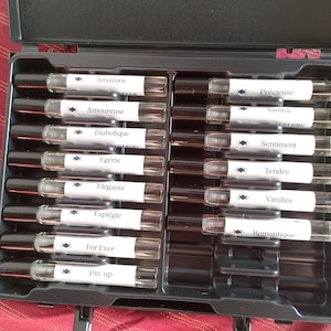 Può includere: Una valigetta nera contenente piccoli flaconi di profumo. Ogni flacone ha un tappo nero e un'etichetta con un nome unico. Le etichette includono nomi come "Amazone", "Amoureuse" e "Diabolique". I flaconi sono disposti in file all'interno della valigetta.