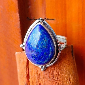 Puede incluir: Un anillo de plata con una piedra de lapislázuli en forma de lágrima. La piedra es de un azul intenso con motas doradas. El anillo tiene una banda sencilla y está adornado con pequeñas cuentas de plata.