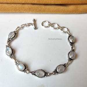 May include: A silver bracelet featuring oval-shaped moonstone cabochons. The bracelet has a toggle clasp and is linked with a series of small, decorative links. The moonstones are set in ornate silver bezels. The text "NewJewelryArtisans" is visible.