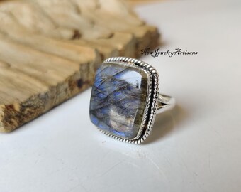 Può includere: Un anello in labradorite a taglio quadrato con una fascia in argento. La gemma mostra sfumature blu e grigie. L'anello ha un design a corda intrecciata attorno alla pietra. Il testo "New Jewelry Artisans" è visibile.