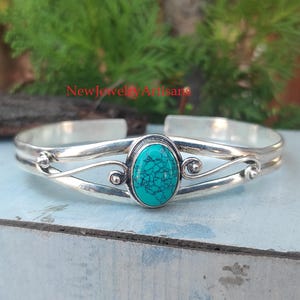 May include: Silver cuff bracelet featuring a turquoise stone set in a swirl design. The stone is oval shaped and has a mottled pattern.