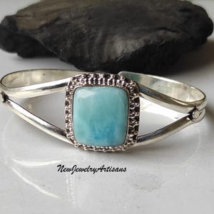 May include: A silver cuff bracelet with a square turquoise blue gemstone set in a silver bezel. The bracelet has a simple design with a curved shape.