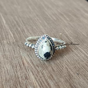 Peut inclure: Bague en argent ornée d'une pierre en forme de goutte d'eau. La pierre est principalement blanche avec des taches noires, encadrée d'un motif torsadé. L'anneau présente de petits détails perlés. Le texte "New Jewelry Artisans" est visible.