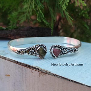 May include: A silver bangle bracelet with two raw gemstones set into the metal. The stones are a green and a pink color.