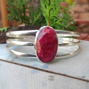 May include: A silver bangle bracelet with a large, oval-shaped, red gemstone set in the center. The bracelet has three bands.