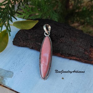 Op de afbeelding: Een zilveren hanger met een roze ovale edelsteen. De hanger heeft een delicate zilveren rand en is geplaatst tegen een houten achtergrond.