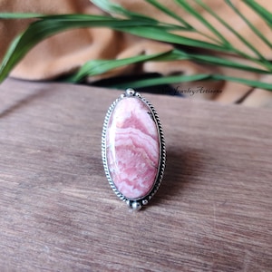 Peut inclure: Bague ovale avec une grosse pierre rose et blanche et une bordure argentée texturée. La bague a un motif décoratif en haut et en bas. La bague est posée sur une surface en bois.