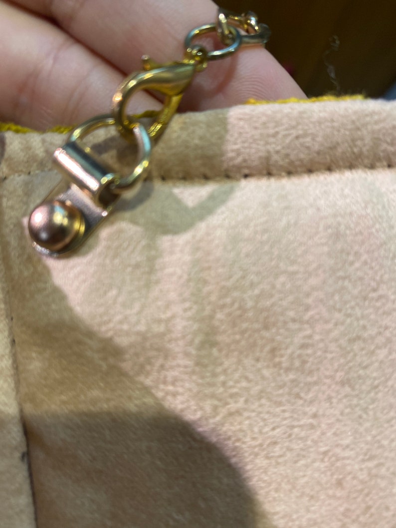 K&ouml;nnte beinhalten: Eine goldfarbene Kette mit einem Verschluss, der an einer beigefarbenen Stoffoberfl&auml;che befestigt ist.