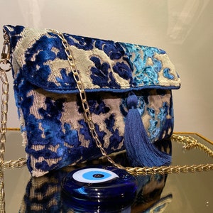Op de afbeelding: Een blauwe en grijze velvet clutch met een gouden kettingband en een blauwe kwast. De tas heeft een bloemenpatroon en een blauwe kwade oog charme.