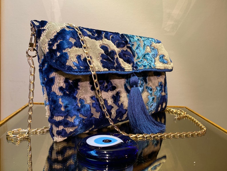 Op de afbeelding: Een blauwe en grijze velvet clutch met een gouden kettingband en een blauwe kwast. De tas heeft een bloemenpatroon en een blauwe kwade oog charme.