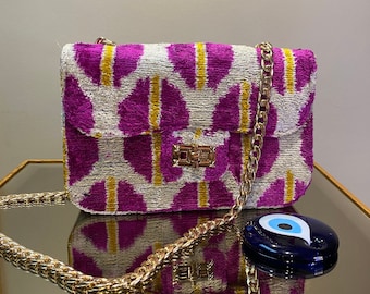 May include: A pink and white patterned velvet shoulder bag with a gold chain strap and a gold clasp. The bag is decorated with a blue evil eye charm.