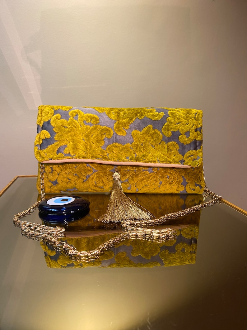 K&ouml;nnte beinhalten: Eine gelbe und graue Clutch aus Samt mit einer goldenen Kettenkette und einem goldenen Quasten. Die Tasche hat einen blauen b&ouml;sen Blick-Anh&auml;nger an der Kette.