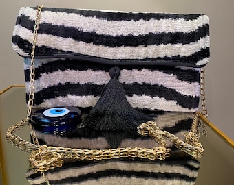 Puede incluir: Un bolso de mano de estampado de cebra en blanco y negro con un borla negra y un amuleto de ojo azul. El bolso tiene una correa de cadena dorada.